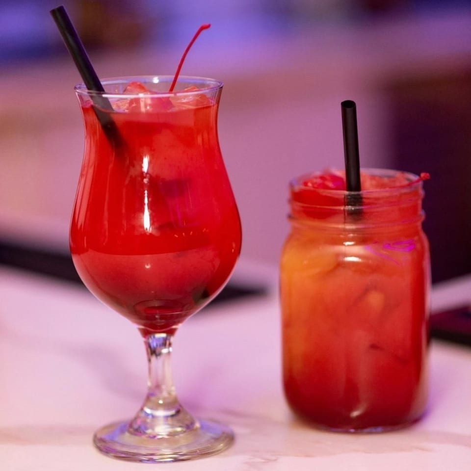 Two vibrant red cocktails, one in a hurricane glass and one in a mason jar, on a table.
