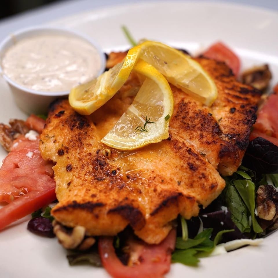 Grilled salmon with lemon slices, mixed greens, tomatoes, and a side of dressing.