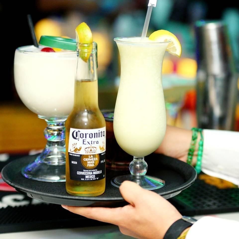 A server carries a tray with two creamy cocktails and a Coronita beer bottle.