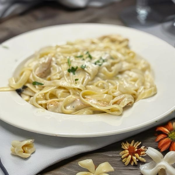 Creamy fettuccine alfredo with parsley on a white plate, next to decorative flowers.