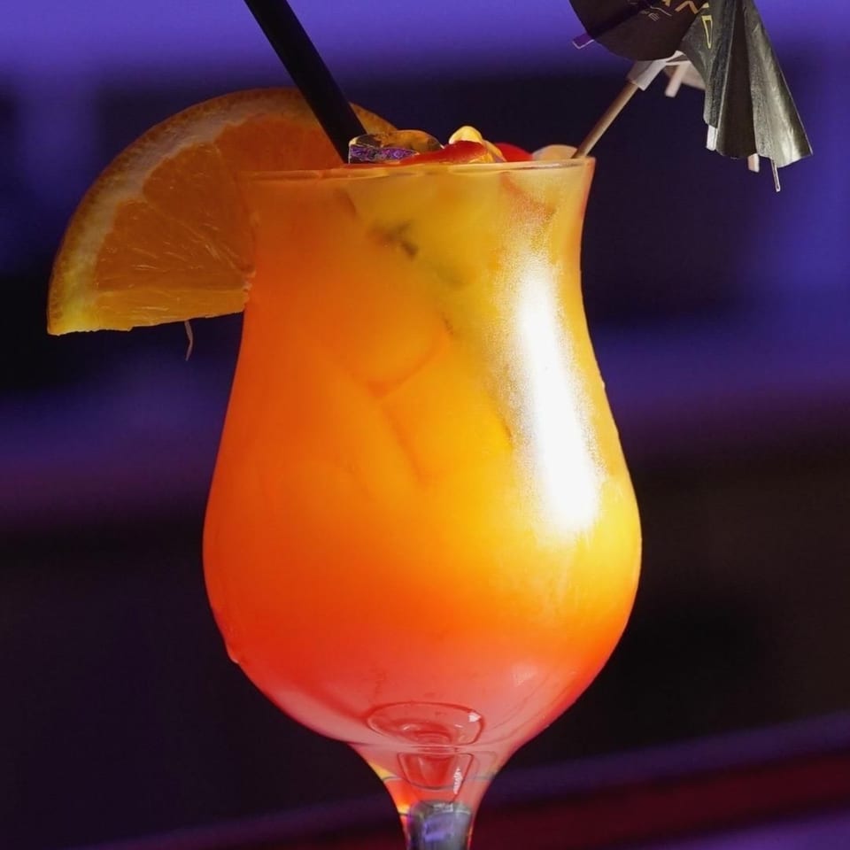 A vibrant orange and red cocktail, garnished with an orange slice and a small umbrella.