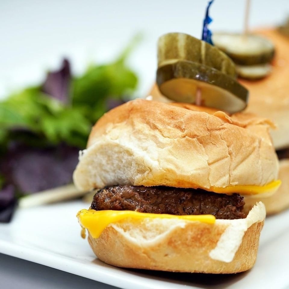 Close-up of a Jimmy's Grand Cafe mini cheeseburger slider with a pickle spear on a white plate.