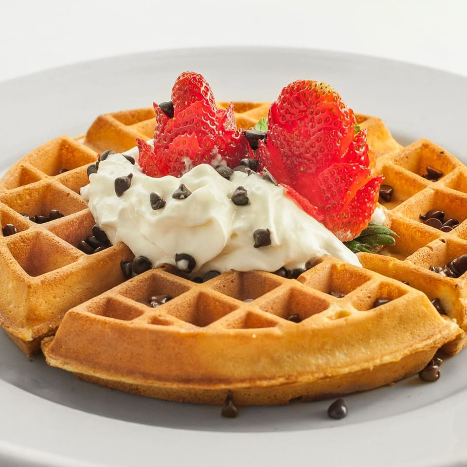 Golden waffle topped with whipped cream, chocolate chips, and fresh strawberries on a white plate.