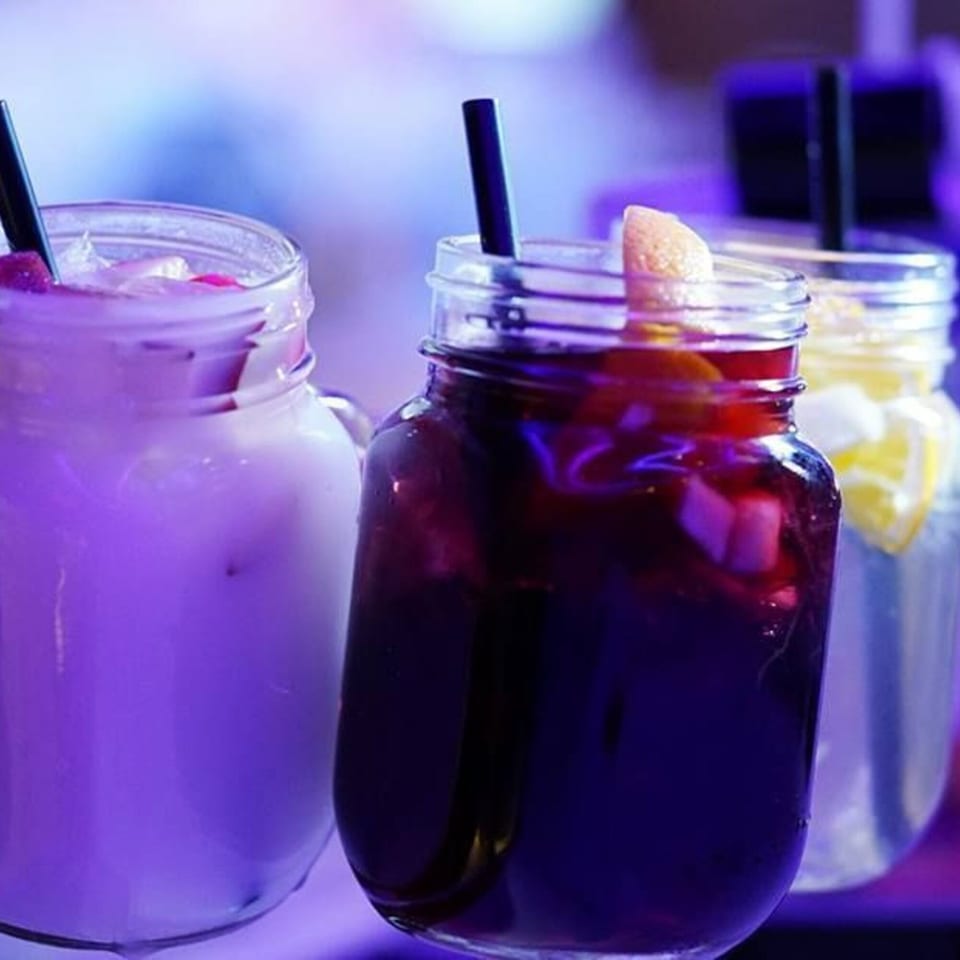 Three colorful drinks in mason jars with straws, one purple, one dark red, and one yellow.