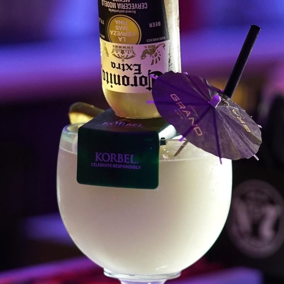 A Corona Extra bottle inverted into a white margarita with a Korbel tag and a purple umbrella.